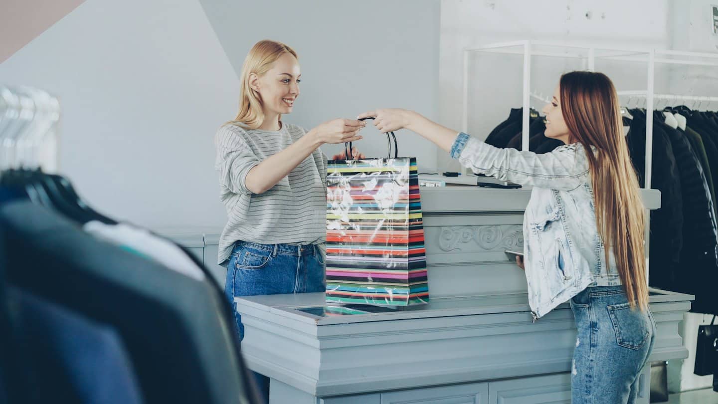Dependiente entrega bolsa de compras colorida a cliente en tienda de ropa.