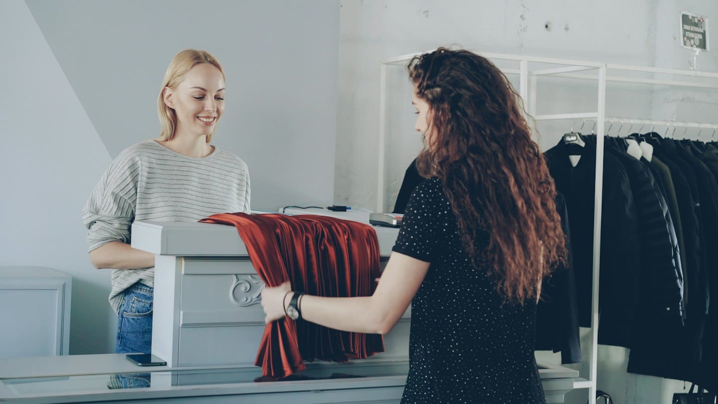 Cliente muestra prenda roja a dependienta sonriente en el mostrador.